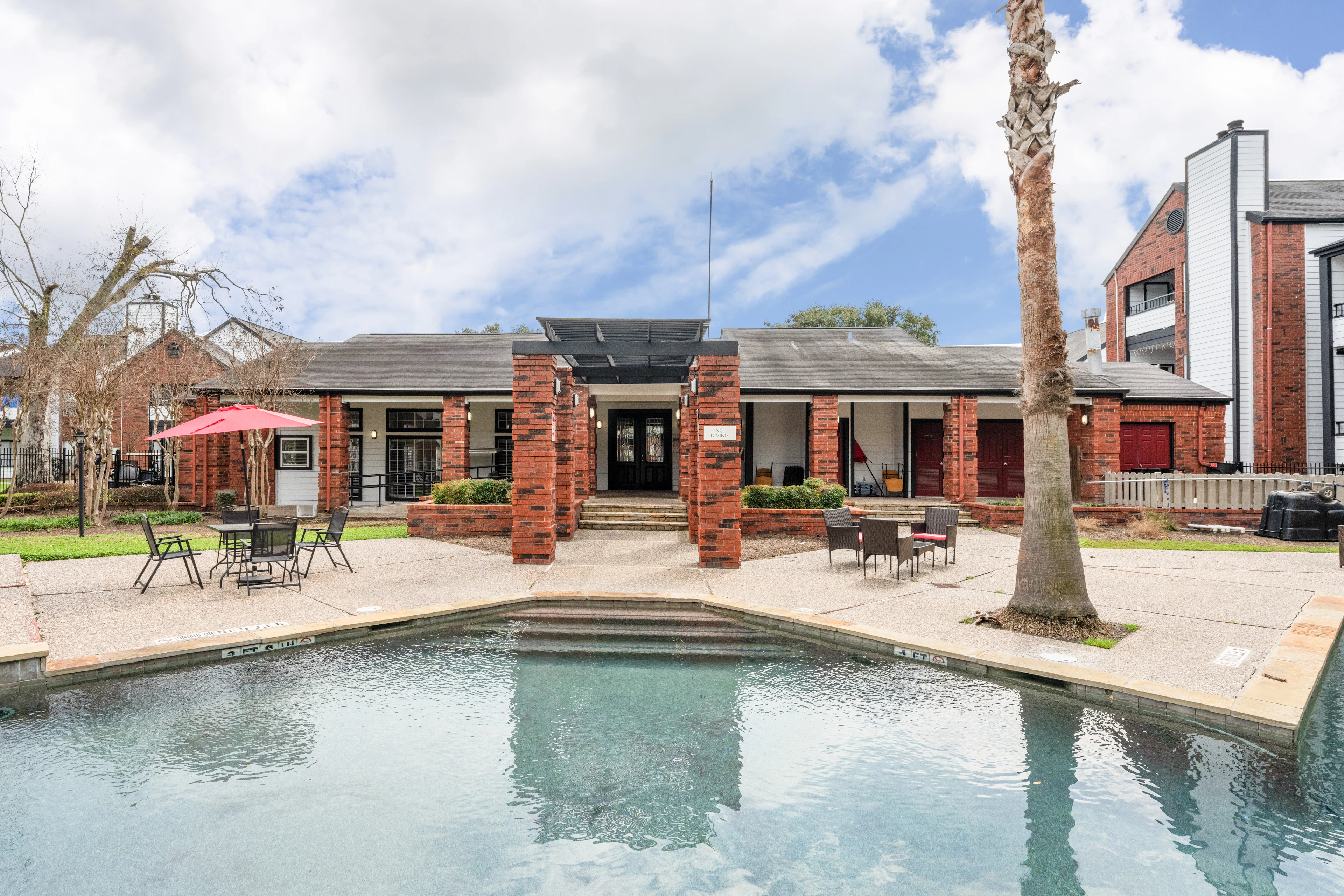 Alberca exterior y clubhouse de ladrillo con porche cubierto y palmeras en West Oaks Landing, en el oeste de Houston.